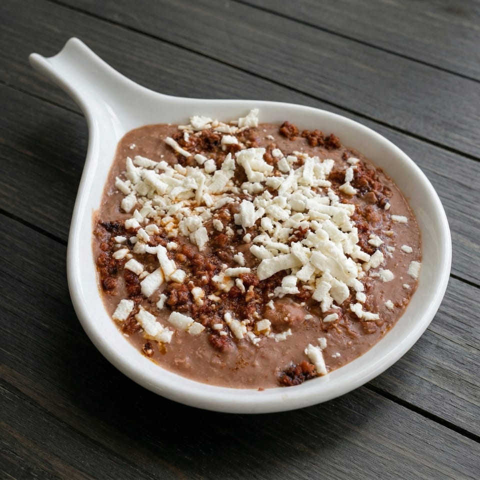 Refried beans topped with crumbled chorizo and white cheese in a white serving dish on a dark wooden table.