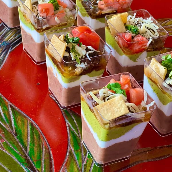 Seven layered dip cups with guacamole, beans, cheese, tomatoes, and tortilla chips on a red table.