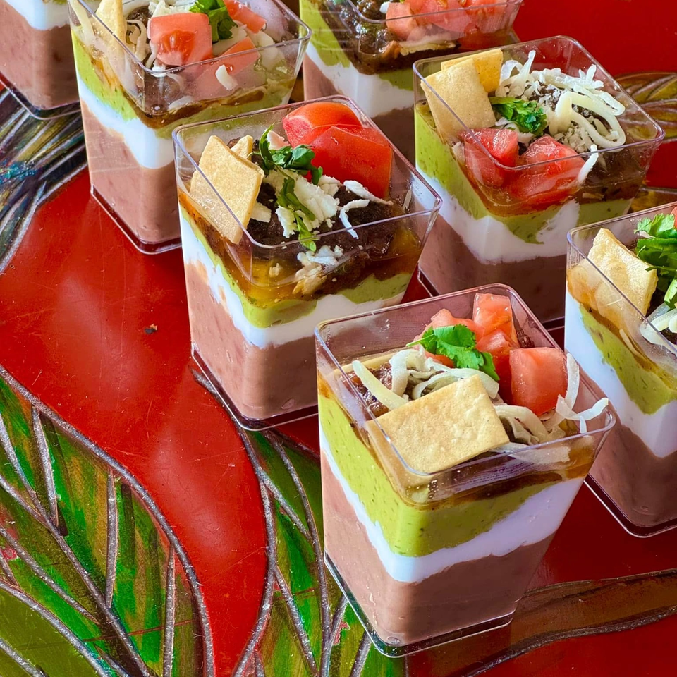 Seven layered dip cups with guacamole, beans, cheese, tomatoes, and tortilla chips on a red table.