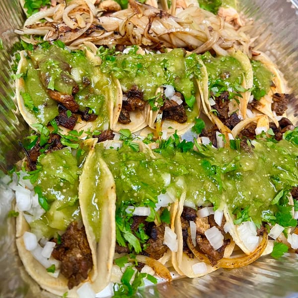 Close-up of several street tacos with meat, onions, cilantro, and green salsa in a foil tray.